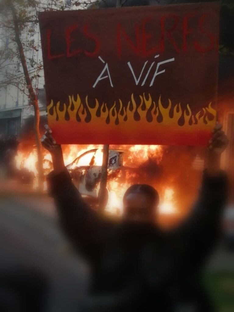 Photo de manif : une femme brandit une pancarte "Les nerfs à vif". Derrière elle on voit une voiture en feu.