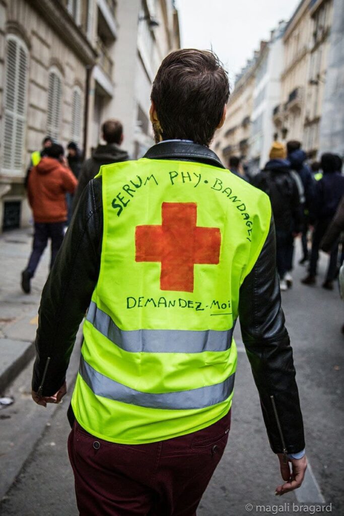 Photo de Magali Bragard. Photo d'un street-medic vu de dos en manif. Sur son Gilet Jaune : croix rouge, "sérum phy, bandages. Demandez-moi".