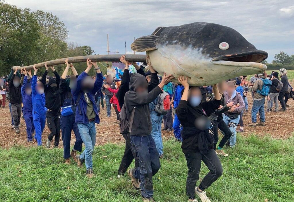Photo des Soulèvements de la Terre. A Sainte-Soline les manifestants portent une canalisation en direction du camp, un gros poisson (mascotte) posé dessus 