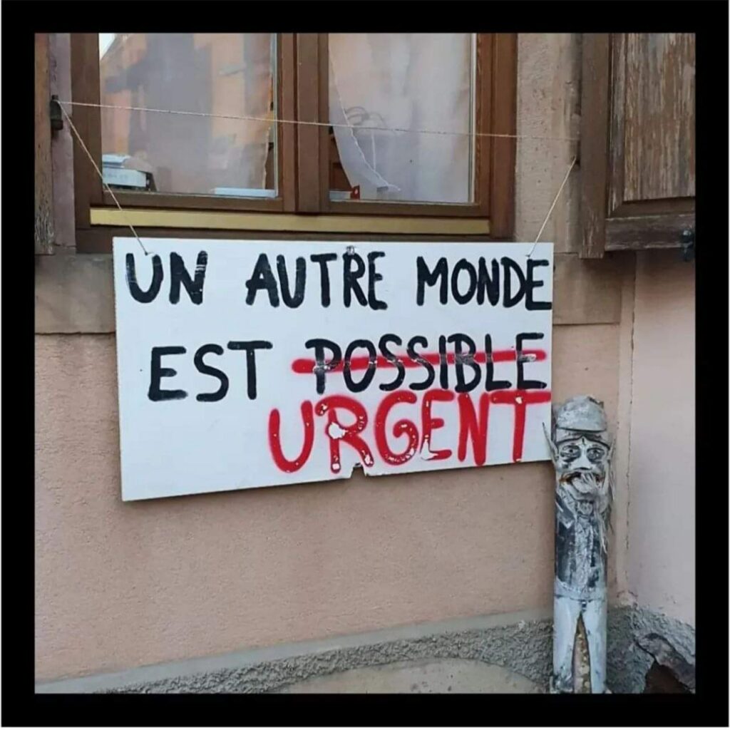 Pancarte accrochée à une fenêtre : "Un autre monde est possible." Le mot "possible" a été rayé et remplacé, en rouge, par "urgent".