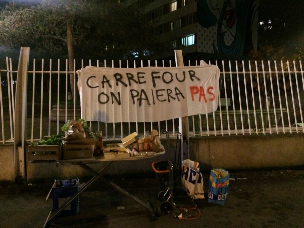 Photo du collectif Carrefour Retire Ta Plainte : 
En extérieur, table avec nourriture et banderole "Carrefour, on paiera pas" 