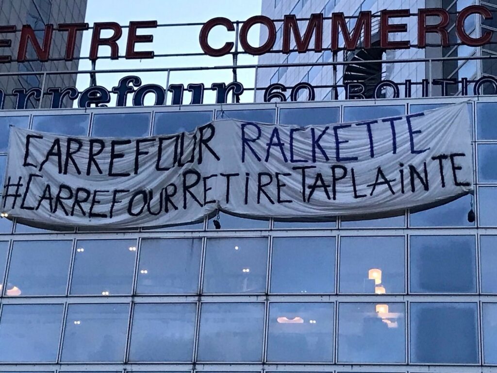 Photo du collectif Carrefour Retire Ta Plainte : 
Devanture d'un magasin Carrefour, grande banderole "Carrefour rackette ! #CarrefourRetireTaPlainte""