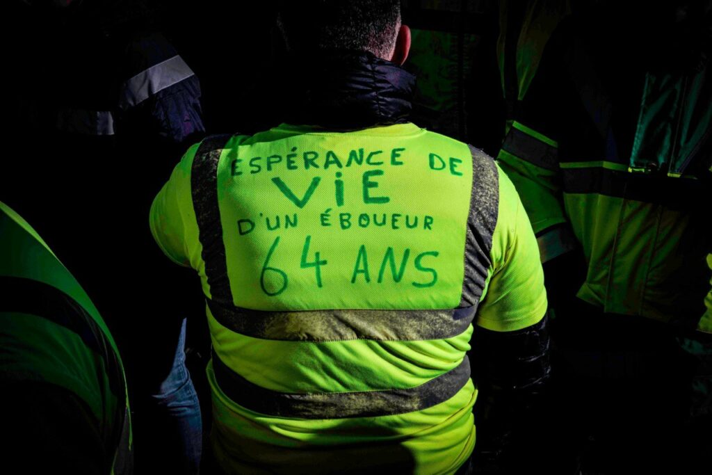 Photo d'un homme de dos, en blouson de travail fluorescent sur lequel est écrit : "Espérance de vie d'un éboueur : 64 ans".