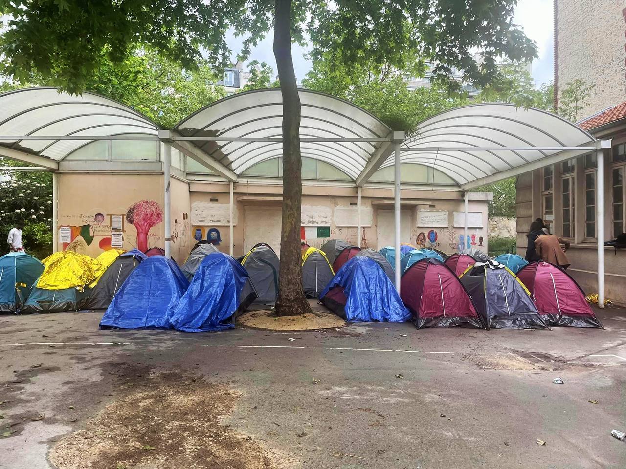 Photo crédit inconnu. 
On voit une vingtaine de tentes sous le préau d'une école. 