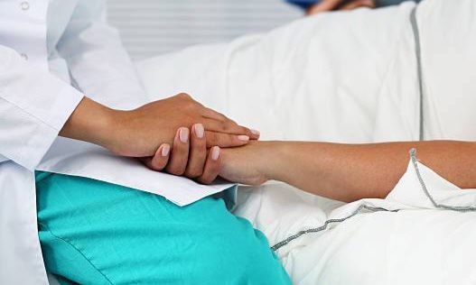 Photo. On voit une soignante assises sur le bord du lit d'un ou une malade alité·e, elle lui tient la main. 