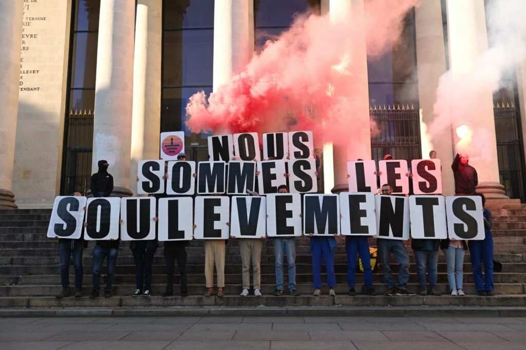 Photo crédit inconnu. Un groupe de personnes sur des marches, avec des fumigènes rouges et tenant des lettres formant le message : "Nous sommes les soulèvements ". 