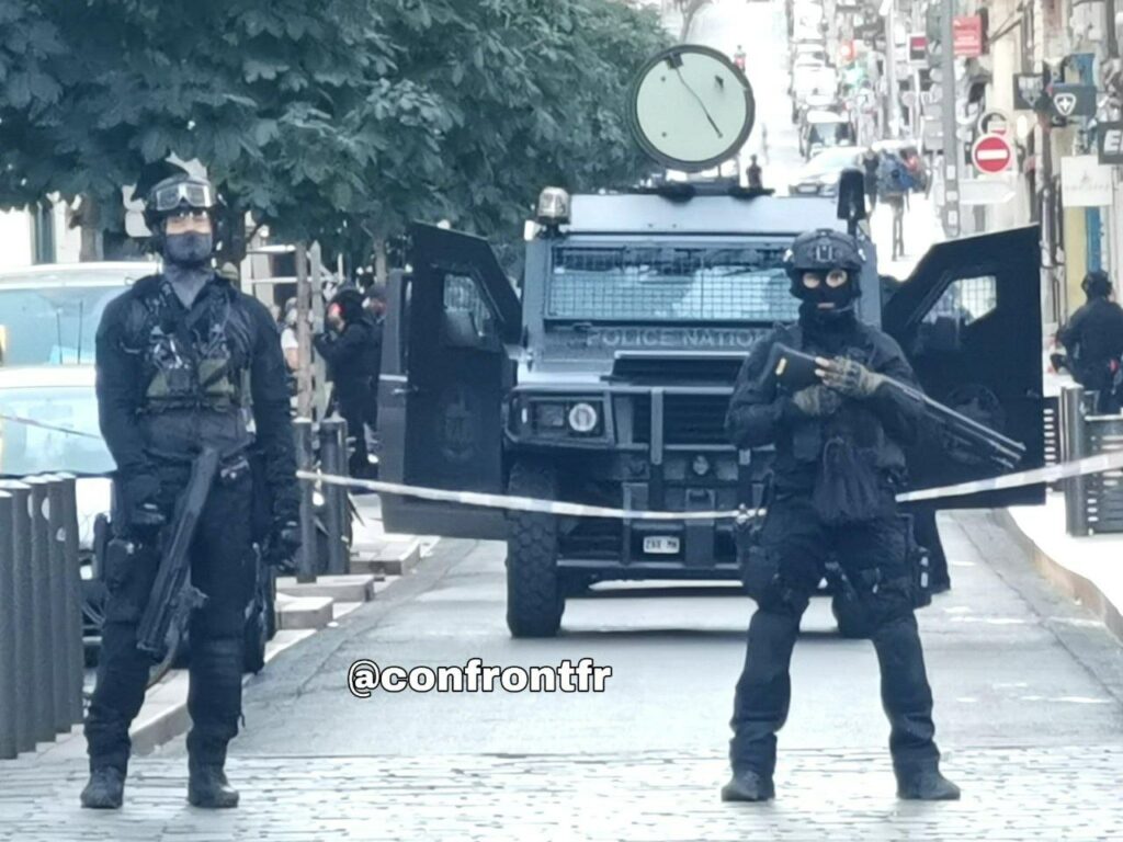 Photo crédit @confrontfr. 
Photo prise à Marseille le 1er juillet 2023. 2 policiers du RAID, tout équipés bloquent une rue et regardent fixement le photographe. Derrière eux de la ribalise a été déployée devant leur véhicule. 
