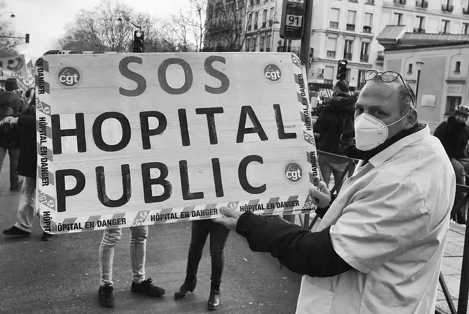 Photo crédit inconnu : un soignant porte une pancarte sur laquelle il est noté : "SOS hôpital public", "hôpital en danger". On voit des tickets de la CGT. 