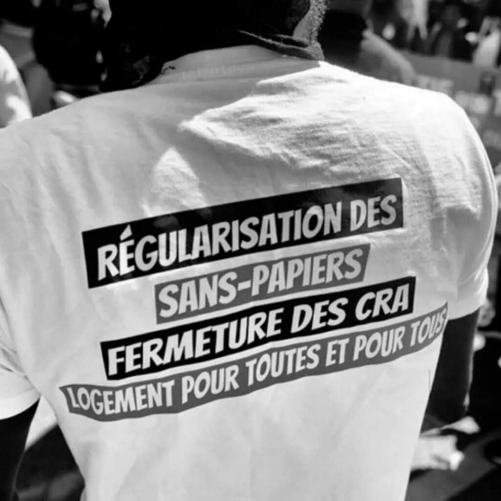 Photo en noir et blanc. Dos du tshirt d'une personne sur lequel il est écrit : "Régularisation des sans-papiers, fermeture des CRA, logement pour toutes et tous"