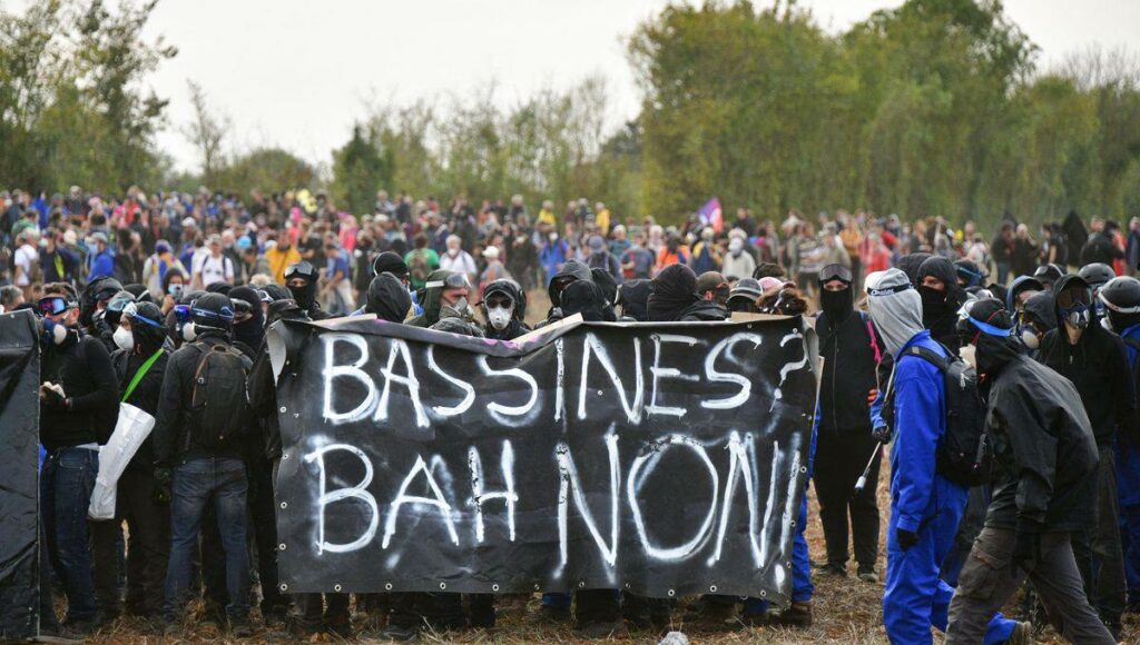 Photo de Pascal Lachenaud, prise à Sainte-Soline. On voit de nombreux manifestant·es. Au premier plan, un groupe, vêtu en noir et équipé, porte une grande banderole noire sur laquelle il est écrit "Bassines ? Bah non ". 