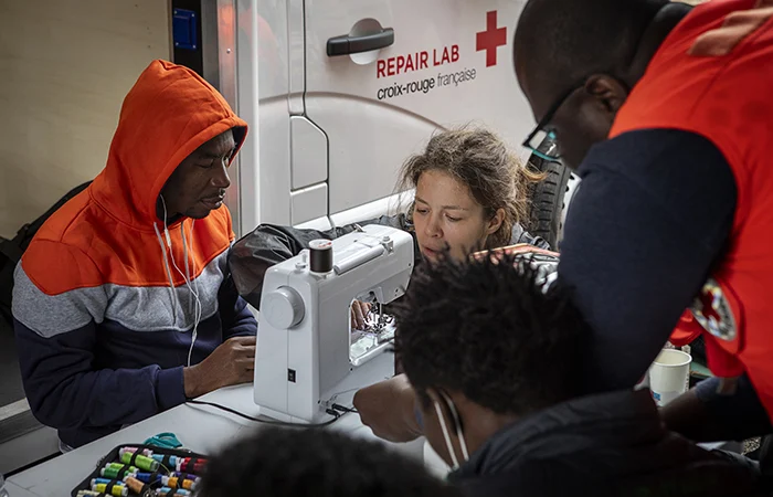 Photo de la Croix-Rouge Française où on voit une femme montrer à plusieurs hommes comment se servir d'une machine à coudre. En arrière plan, on voit le camion de la Croix-Rouge avec le logo de l'ONG et l'inscription "Repair Lab".