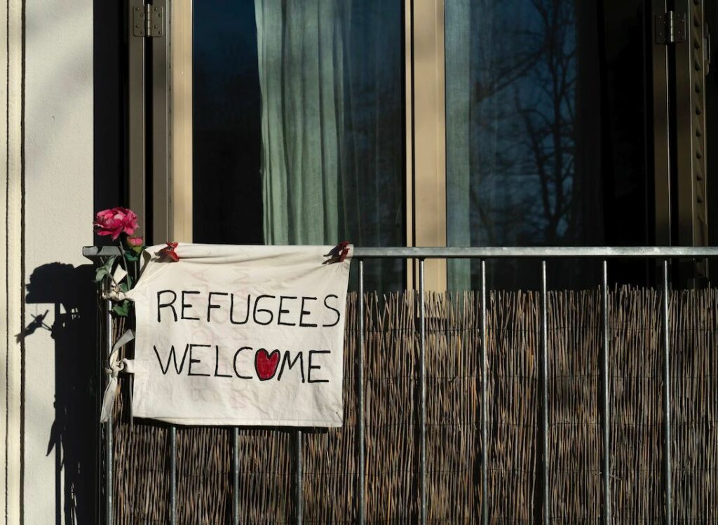 Photo d'une banderole accrochée sur un balcon sur laquelle il est écrit "Refugees welcome", le O a été remplacé par un coeur rouge. Une rose est accrochée à la banderole.