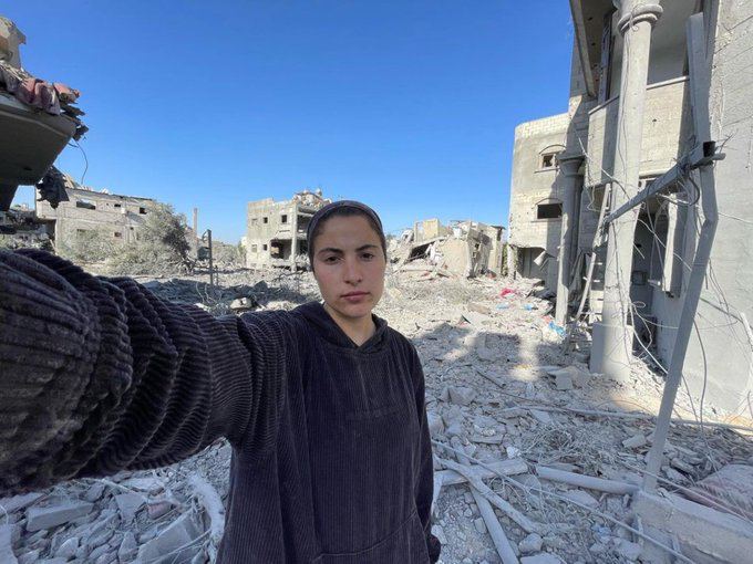 Selfie de Bisan Owda pris le 3 décembre 2023 à Khan Younès. On voit derrière elle un champ de ruine, toutes les habitations ont été détruites, la rue est déserte. 