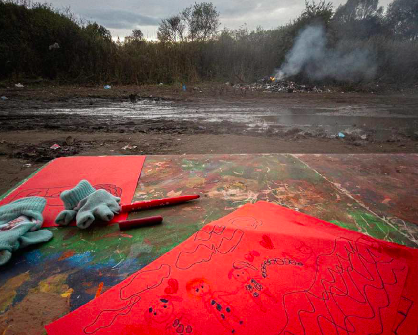 Photo de Luis Lujan. Terrain vague boueux, visiblement en hiver, avec en arrière plan un feu. Au premier plan, on voit un morceau de contreplaqué sur lequel il y a des traces de peinture, deux feuilles de dessin rouges, un feutre et deux gants d'enfant.