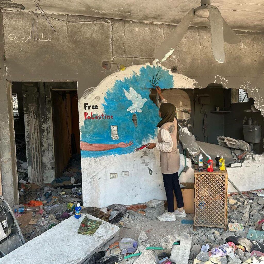 Photo crédit inconnu d'Amal Abu Al-Sabah en train de peindre une fresque sur un mur, on y voit une colombe, une main tendue et le texte "Free Palestine".