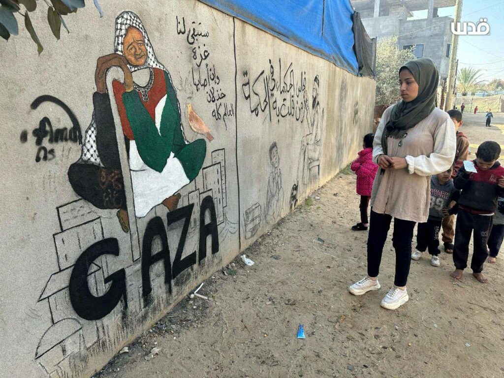 Photo crédit inconnu d'Amal Abu Al-Sabah face à un mur sur lequel elle a peint une vieille femme revêtue du drapeau palestinien et sous laquelle il est écrit "Gaza".