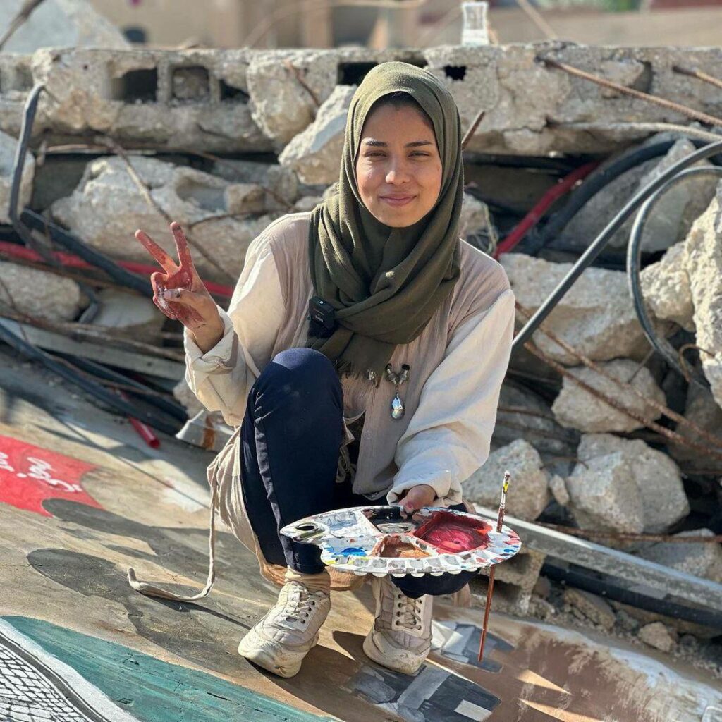 Photo crédit inconnu. Gros plan sur Amal Abu Al-Sabah, souriante, faisant le V de la victoire de sa main droite et tenant de sa main gauche sa palette de couleurs avec laquelle elle peint une fresque. 