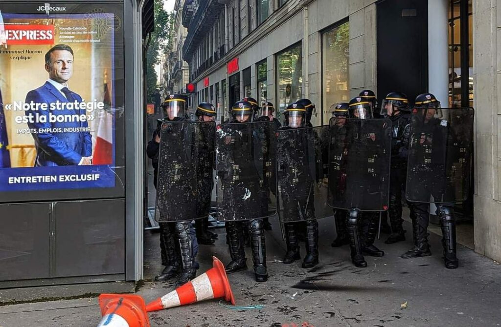 Photo de Bernard Chevalier prise en manif. On voit des CRS derrière leurs boucliers. Juste à leur droite on voit l'affiche d'une une de L'express avec une photo de Macron "Notre stratégie est la bonne". 
