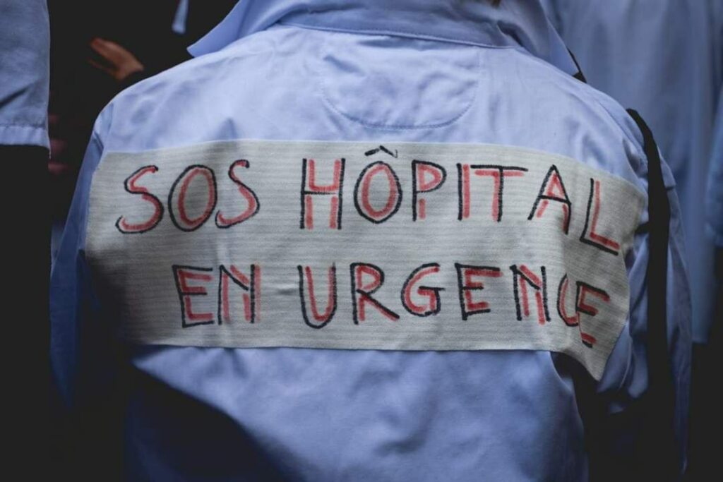 Blouse blanche vue de dos en manif. Message collé sur le dos : "SOS Hôpital en urgence".