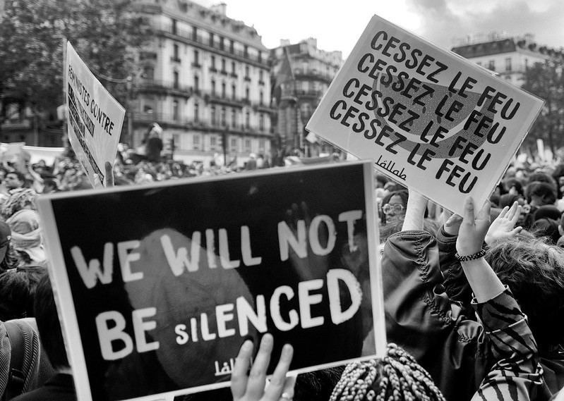 Photo en noir et blanc d'une manif. Au premier plan, une pancarte : "We will not be silenced" (nous ne serons pas silenciés) et un peu en arrière une autre pancarte sur laquelle il est noté 4 fois "Cessez le feu".